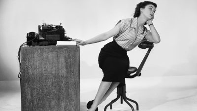 Woman leaning on a desk with a typewriter, looking up thoughtfully, a pencil in hand.