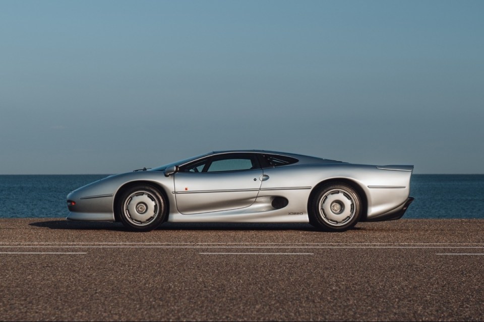Silver Jaguar XJ220 supercar parked on a road next to the ocean under a clear blue sky