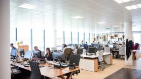 Modern open-plan office space with employees working at computers, bright lighting, and large windows.