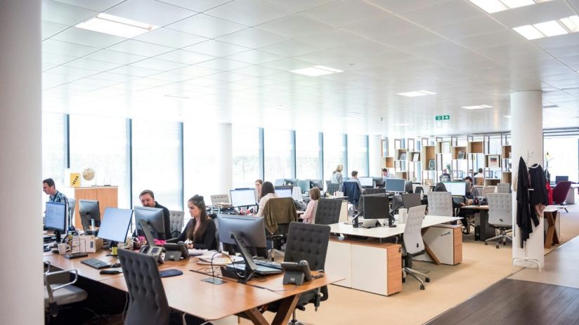 Modern open-plan office space with employees working at computers, bright lighting, and large windows.