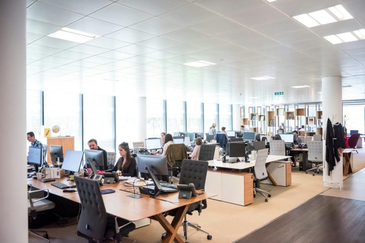 Modern open-plan office space with employees working at computers, bright lighting, and large windows.