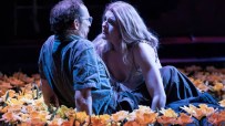 A man and woman in an intimate embrace, surrounded by a field of orange and yellow flowers on a dark stage