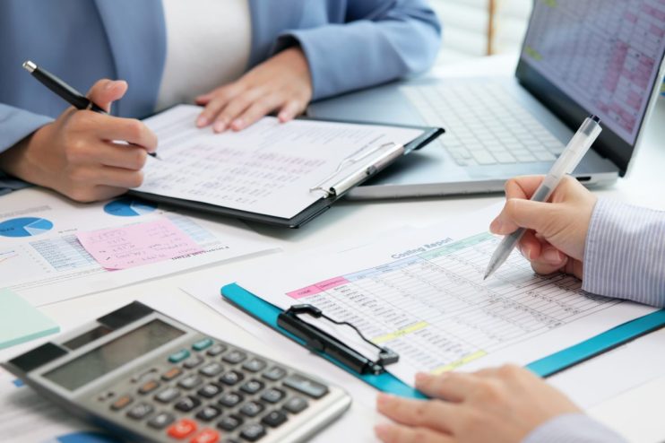 Two business professionals review and analyze a costing report with a calculator and laptop on a desk.