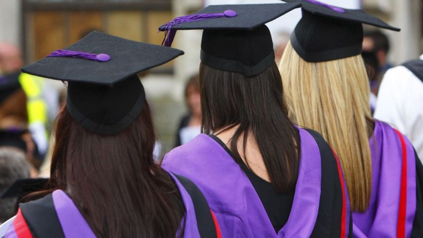Three graduates in black caps with purple tassels and purple academic gowns from behind.