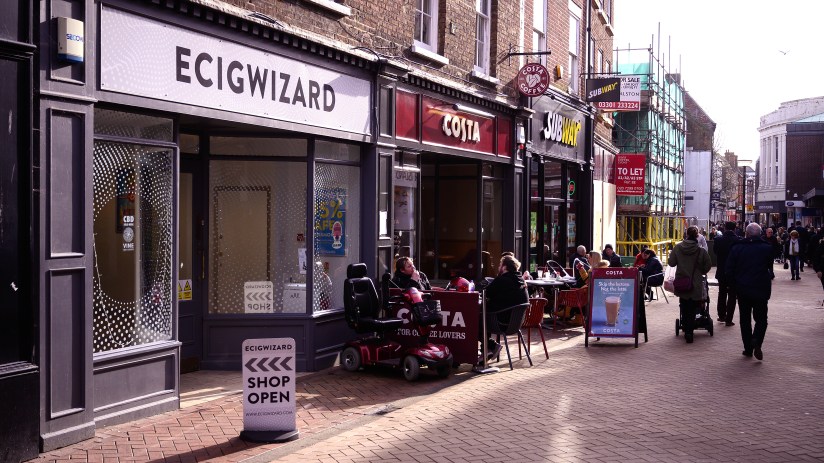 High street vape shop Ecigwizard next to Costa Coffee and Subway, with people walking and sitting outside.