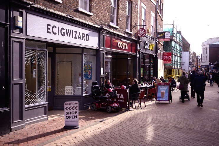 High street vape shop Ecigwizard next to Costa Coffee and Subway, with people walking and sitting outside.
