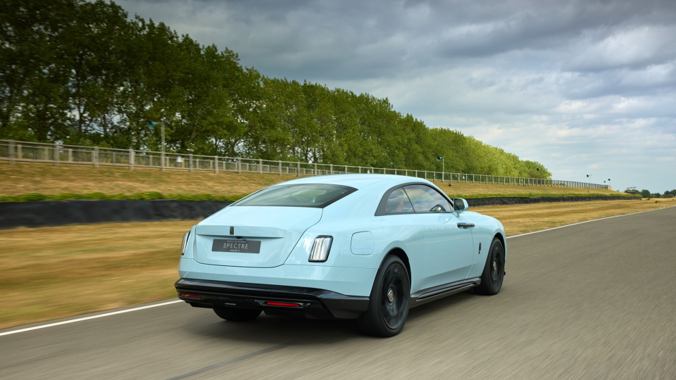 Light blue Rolls-Royce Spectre electric vehicle driving on a race track with motion blur, green trees in background