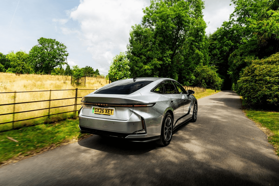 Silver Lexus RZ 350e electric car driving on a paved road through a lush green, tree-lined landscape