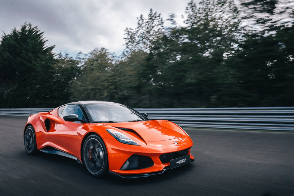 Bright orange sports car, possibly a Lotus Emira, speeding on a race track with blurred trees in background