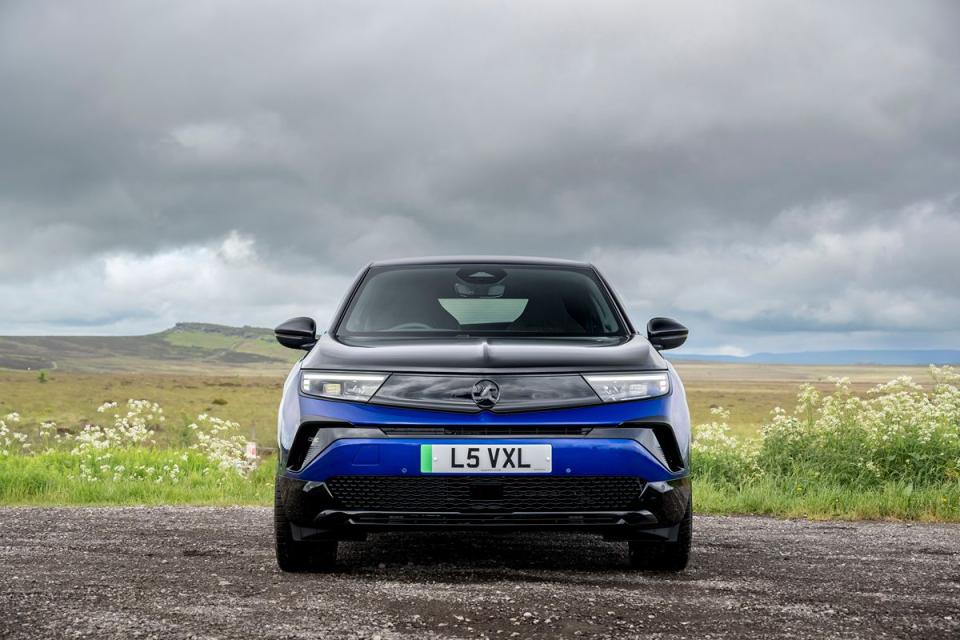 Front view of a blue and black Vauxhall Mokka parked on a gravel road with a cloudy sky and green hills in the background.