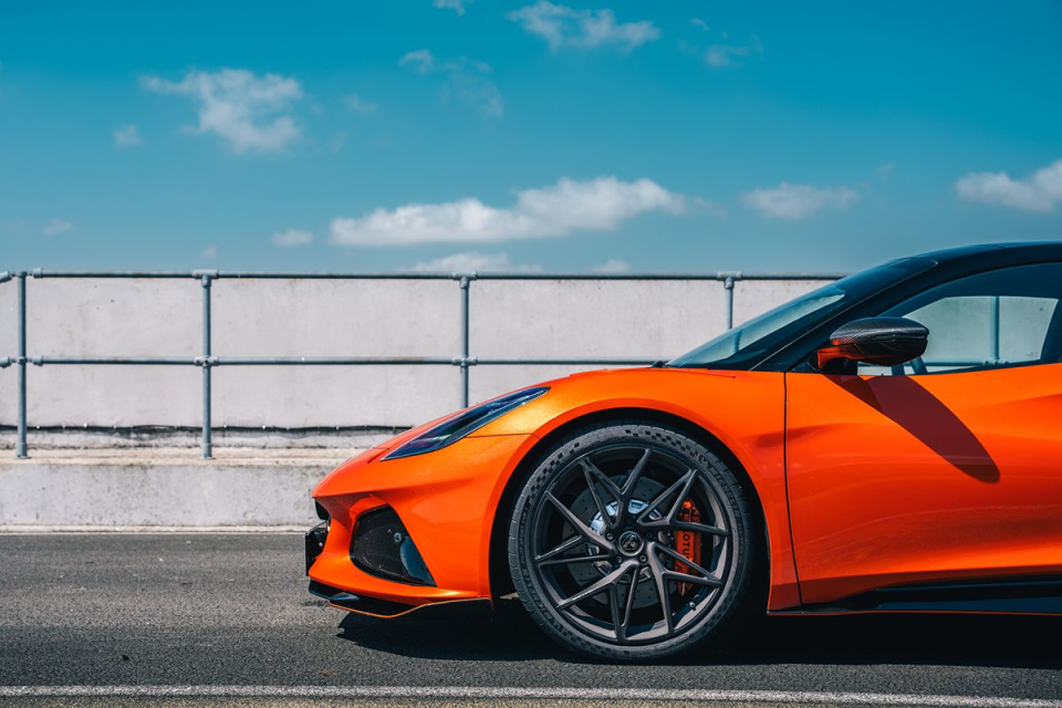 Orange sports car, possibly a Lotus Emira, on an asphalt road under a blue sky with clouds