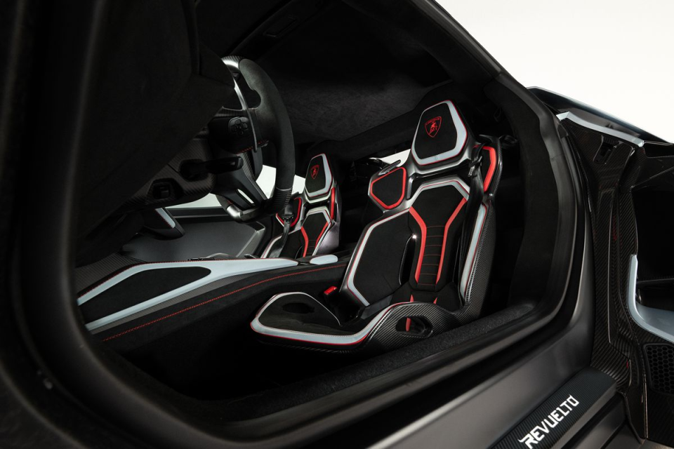 Modern car interior with black and red racing seats, visible steering wheel, and REVUELTO on the sill.