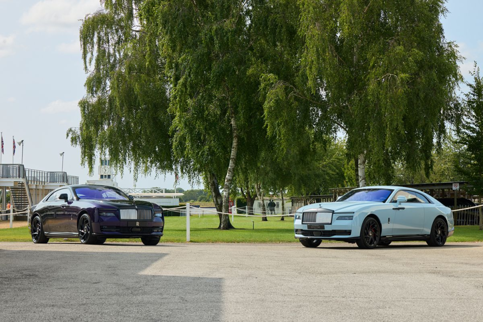 Two luxury Rolls-Royce Spectre electric coupes, one purple and one light blue, parked outdoors under trees.