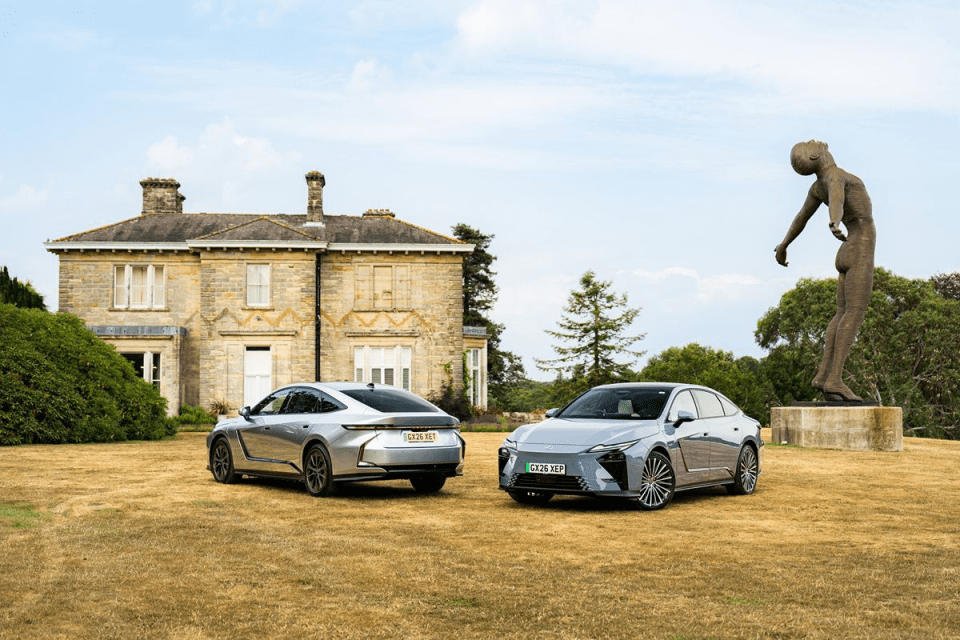 Two sleek electric cars, one silver and one grey, parked on dry grass in front of a stone mansion with a large statue.