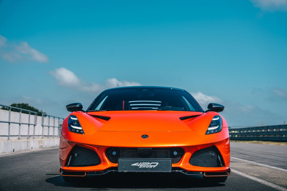 Front view of a bright orange Lotus Emira 420 Sport on a racetrack under a blue sky.