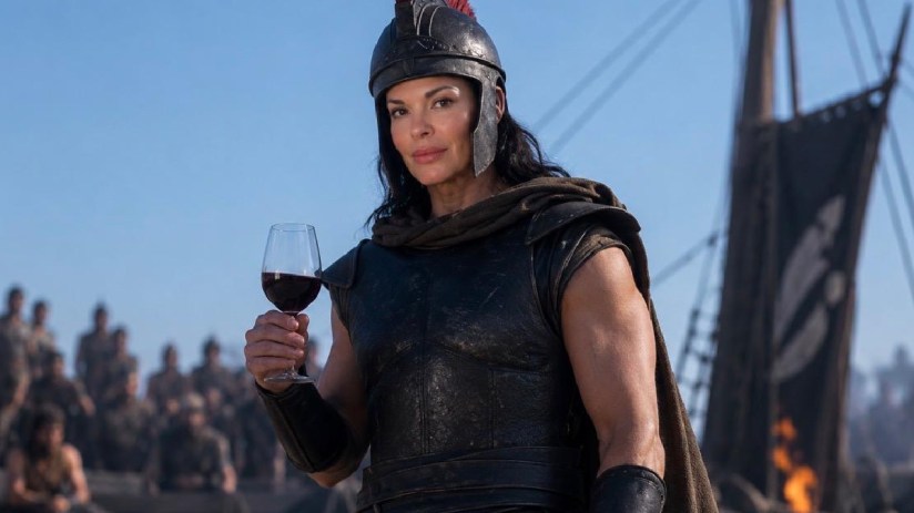 Libby in a warrior costume with a helmet, holding a glass of red wine, with an ancient ship and people in background.