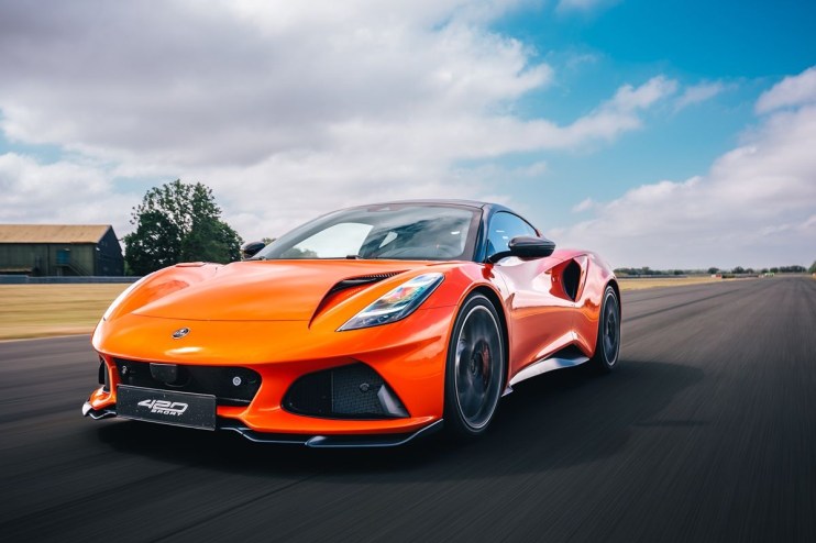 Bright orange Lotus Emira 400 Sport speeding on a racetrack under a blue sky with clouds.