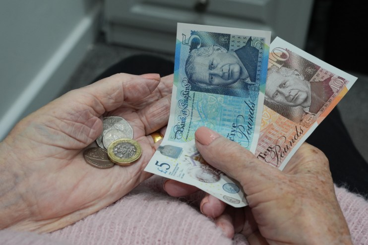 Elderly hands holding British pound notes (&pound;5, &pound;10) and coins, representing pension funds and finances.