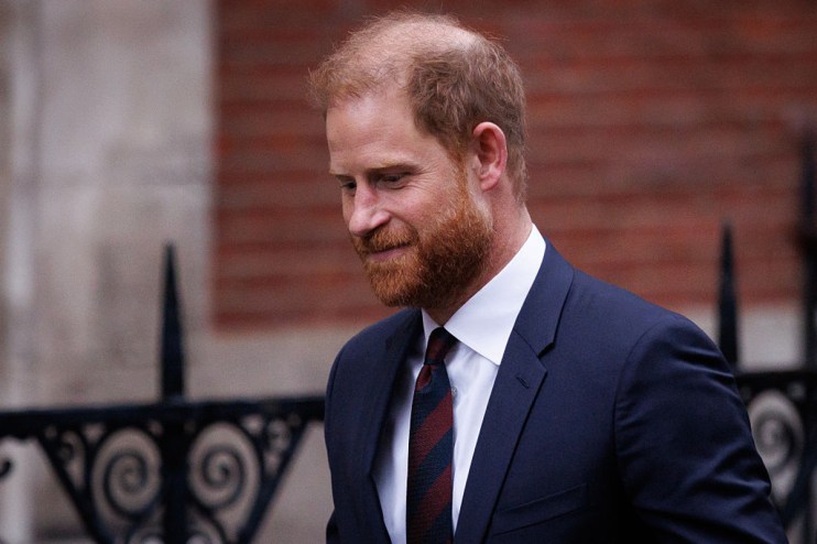 Prince Harry, Duke of Sussex, in a navy suit, white shirt, and striped tie, looking down.