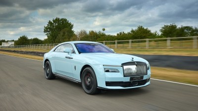 Light blue Rolls-Royce Spectre electric coupe driving fast on a race track under a cloudy sky.