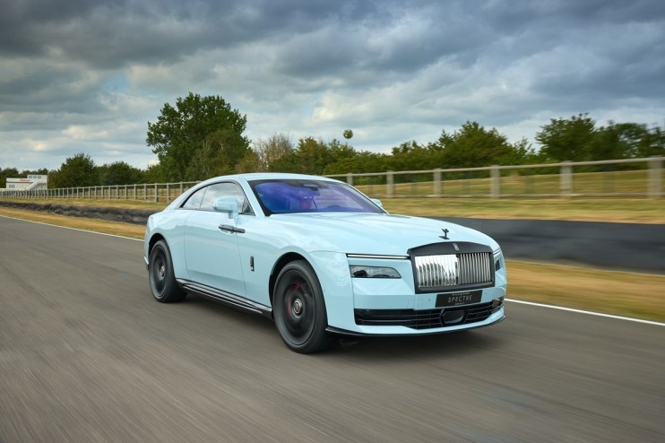 Light blue Rolls-Royce Spectre electric coupe driving fast on a race track under a cloudy sky.