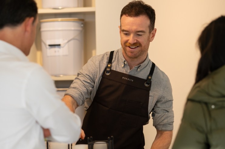 Smiling barista wearing a ROSSLYN apron serving a customer at a coffee shop counter.