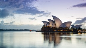 Sydney Opera House at dawn with illuminated sails reflecting on calm water and a cloudy sky