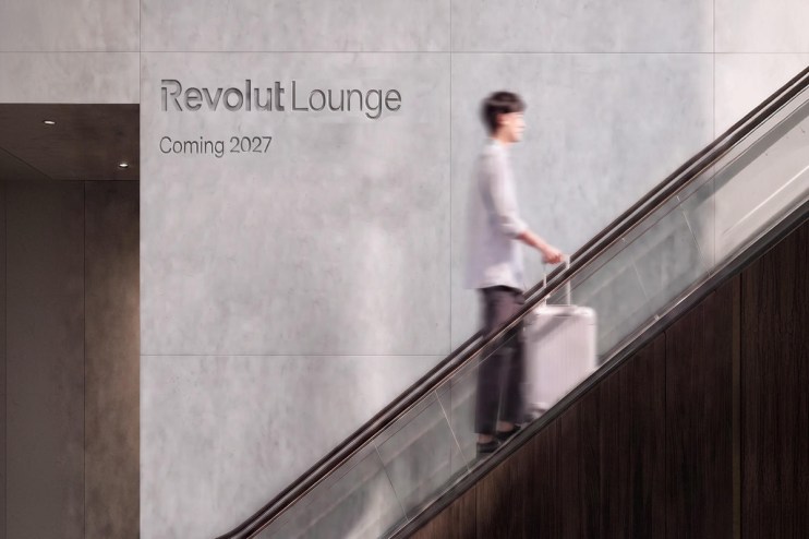 Revolut Lounge sign on a concrete wall with a person on an escalator holding a rolling suitcase.