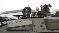 Two people, a woman and a man, standing in the open hatches of a large olive-green military vehicle.