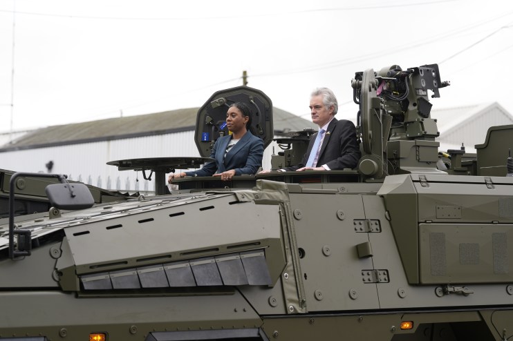 Two people, a woman and a man, standing in the open hatches of a large olive-green military vehicle.