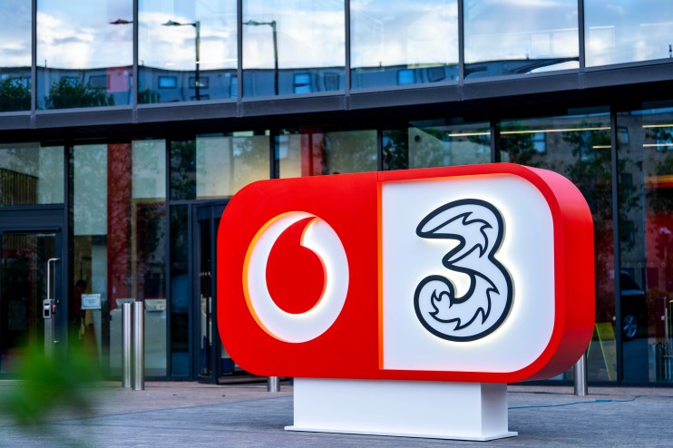 Vodafone and Three company logos on a red and white sign outside a modern glass building