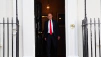 A man in a dark suit and red tie walks through a doorway with black iron railings on either side.