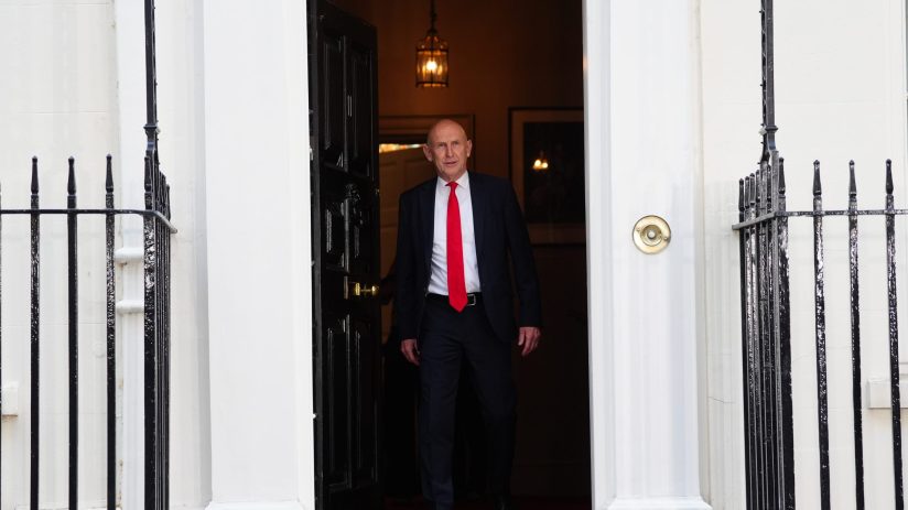 A man in a dark suit and red tie walks through a doorway with black iron railings on either side.