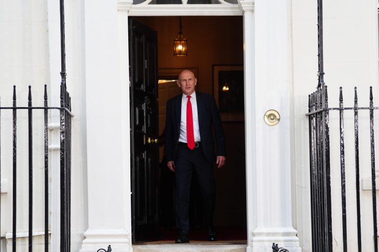A man in a dark suit and red tie walks through a doorway with black iron railings on either side.