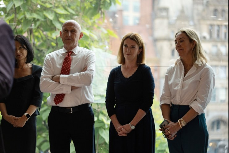 Four professionals, two women and a man, standing indoors with a cityscape visible through a large window.