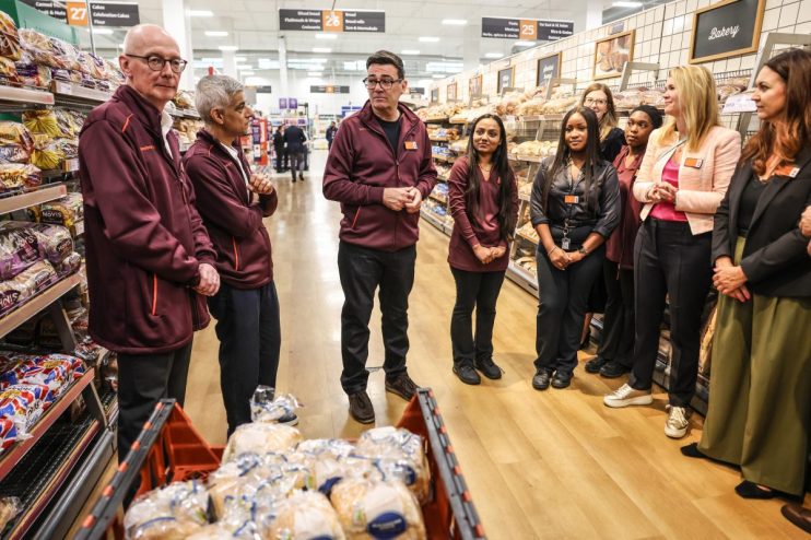 Group of supermarket staff and executives, including Andy Burnham and Sadiq Khan, standing in a bakery aisle.