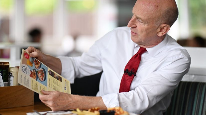 Bald man in white shirt and red tie reading a restaurant menu with a lapel microphone attached