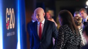 Bald man in a dark suit and red tie walking past a G20 sign with other attendees in background.