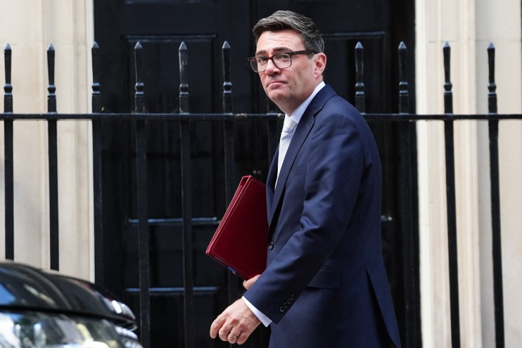 Andy Burnham, Mayor of Greater Manchester, in a suit, holding a red folder, walking past railings