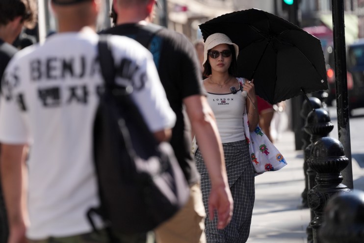 A woman in sunglasses and a hat holding a black umbrella on a sunny city street, with blurred people in the foreground.