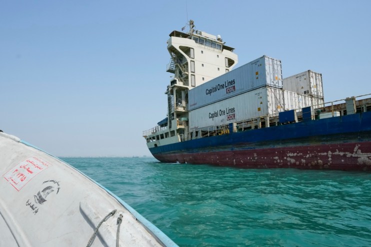 Large cargo ship, Capital One Lines containers visible, on bright blue water, with a small boat in foreground.