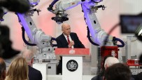 A bald man in a suit speaking at a podium with the Royal Coat of Arms, flanked by two ABB industrial robots.