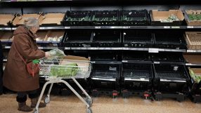 Shopper places produce into a cart in a grocery store aisle with mostly empty shelves, indicating supply shortages.