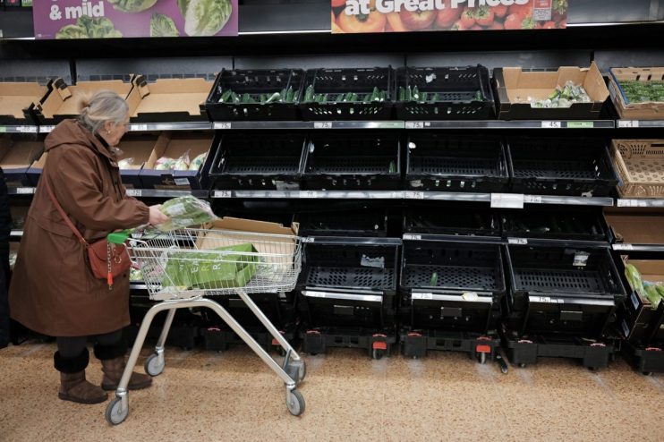 Shopper placing groceries into a cart next to mostly empty supermarket produce shelves, showing limited availability.