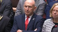 Bald man in a dark suit and red tie, arms crossed, looking intently in a formal setting.