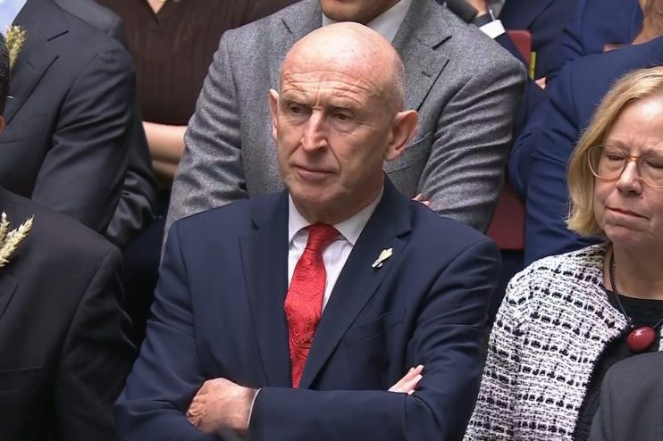 Bald man in a dark suit and red tie, arms crossed, looking intently in a formal setting.