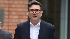 Andy Burnham, Mayor of Greater Manchester, in a dark suit and glasses, walking outdoors with a tote bag.