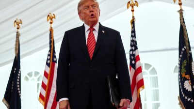 Donald Trump speaking at an event with US and presidential flags, holding a black folder