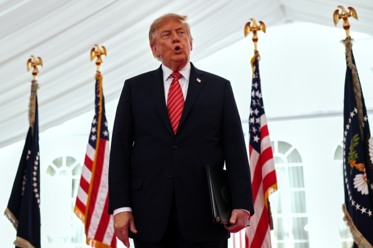 Donald Trump speaking at an event with US and presidential flags, holding a black folder