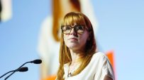 Woman with red hair and glasses at a podium with microphones, a blurred screen in the background.